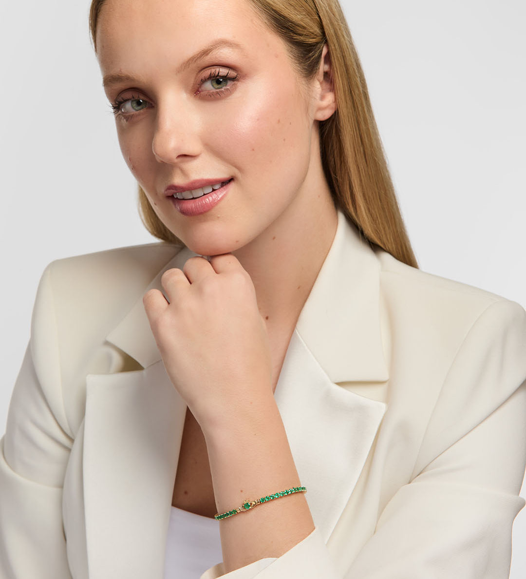 A blonde woman wearing a white blazer and white top, posing with a delicate emerald tennis bracelet featuring Claddagh design on her wrist against a light gray background.