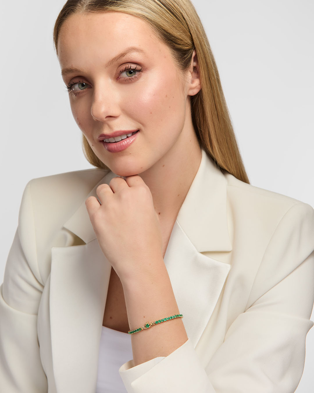 A blonde woman wearing a white blazer and white top, posing with a delicate emerald tennis bracelet featuring Claddagh design on her wrist against a light gray background.