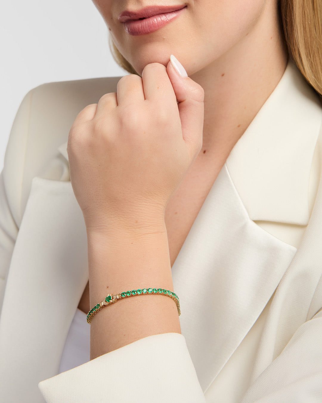A close-up of a woman wearing a delicate gold Claddagh tennis bracelet adorned with green emerald gemstones, paired with a cream-colored blazer.