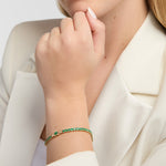 A close-up of a woman wearing a delicate gold Claddagh tennis bracelet adorned with green emerald gemstones, paired with a cream-colored blazer.