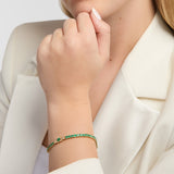 A close-up of a woman wearing a delicate gold Claddagh tennis bracelet adorned with green emerald gemstones, paired with a cream-colored blazer.