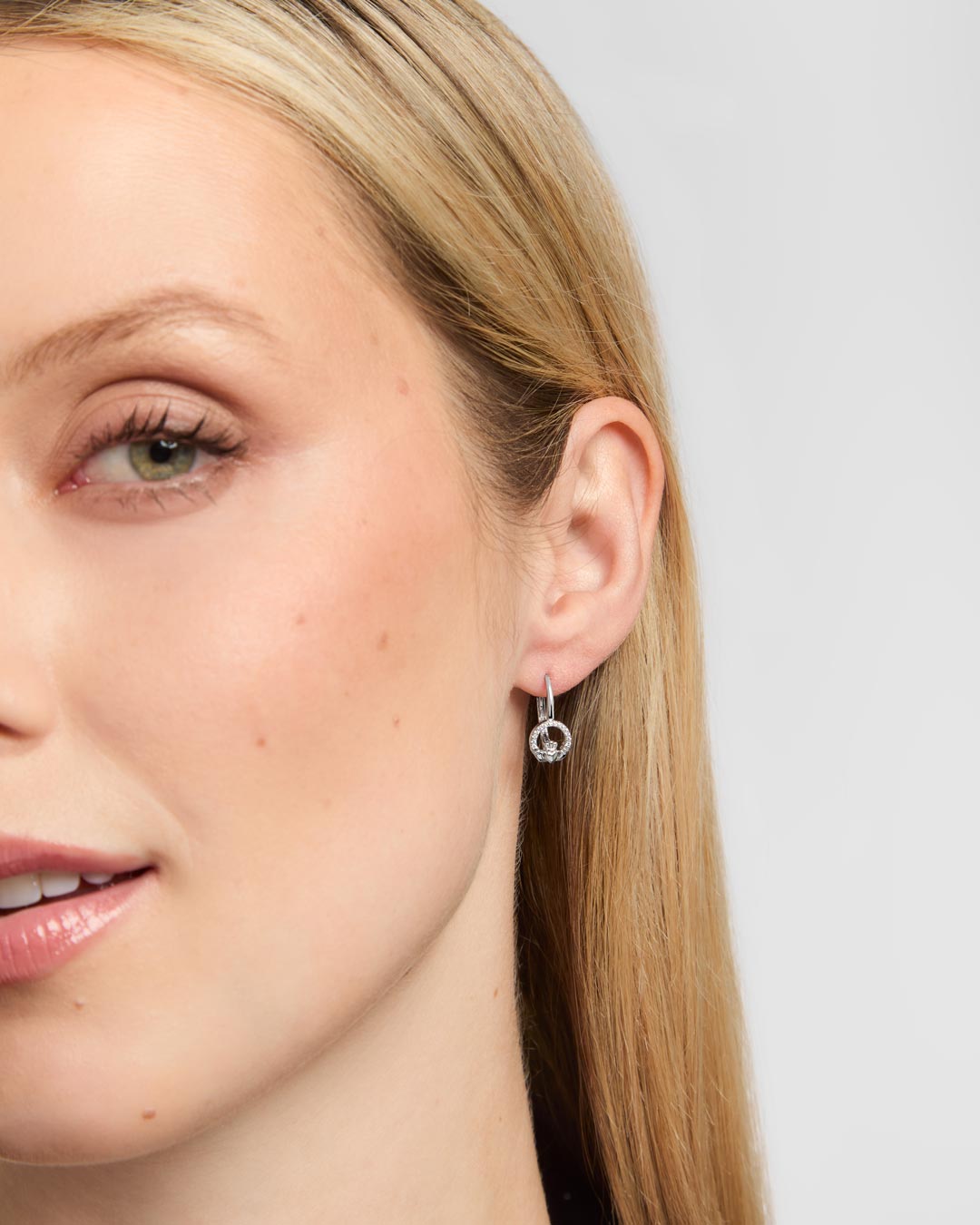Close-up of a woman's ear wearing a delicate silver Claddagh hoop earring with a small dangling circular charm, against a light grey background.