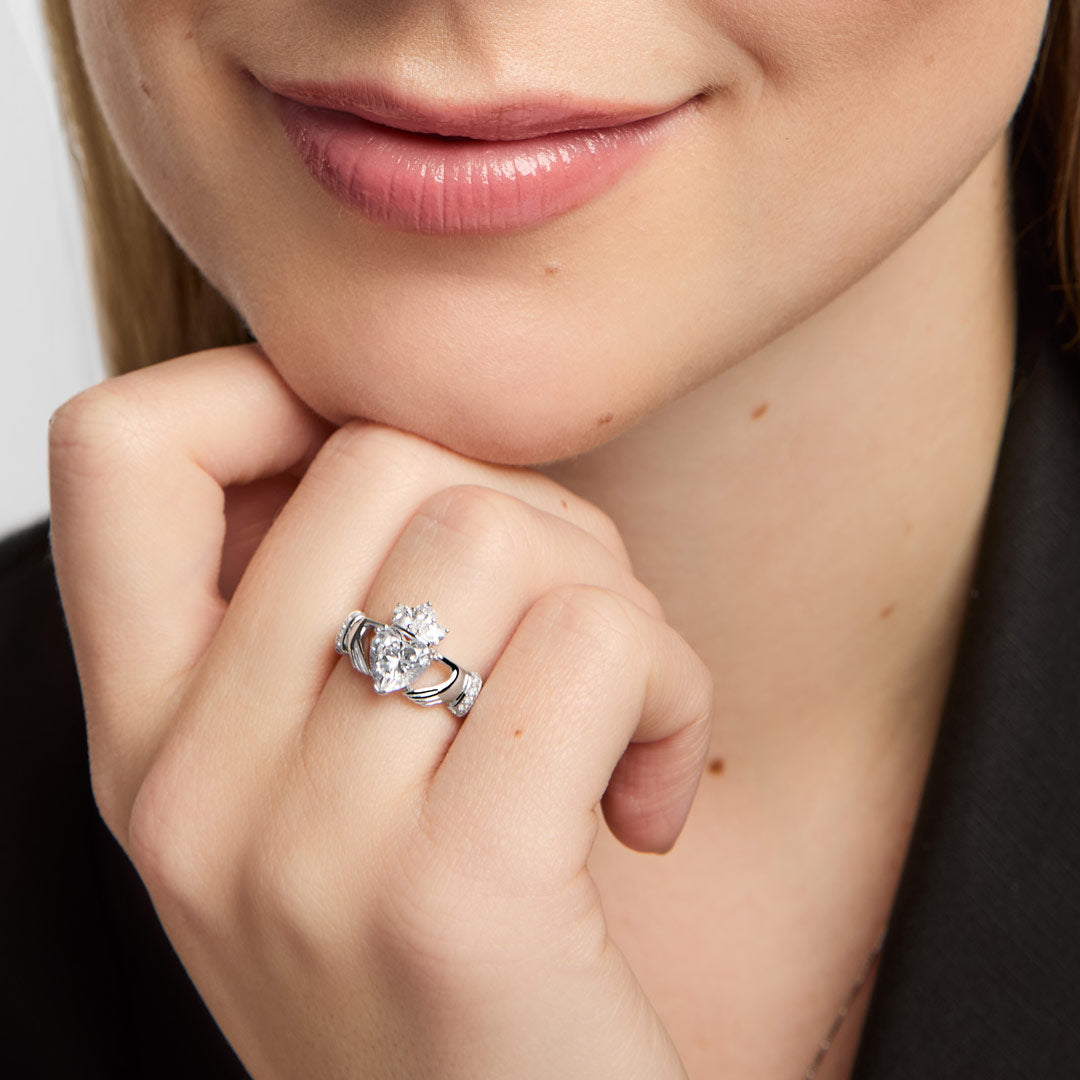 A close-up lifestyle shot of a woman wearing a silver Claddahg ring with a central white stone heart, resting her chin on her hand.