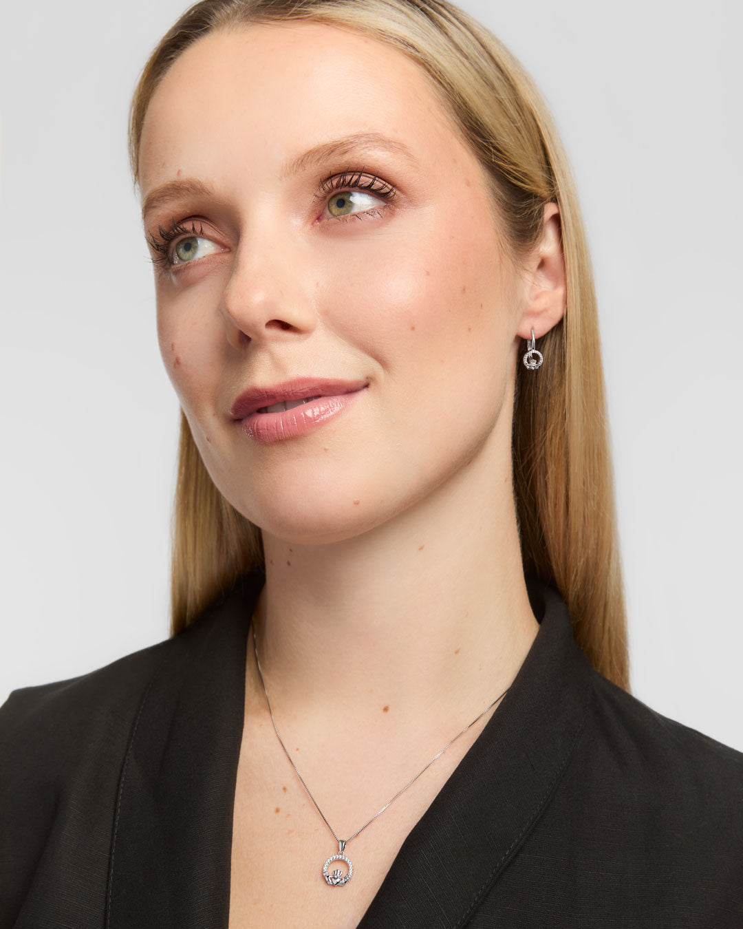 A close-up portrait of a woman wearing a delicate silver Claddagh pendant necklace and matching hoop earrings against a light grey background.