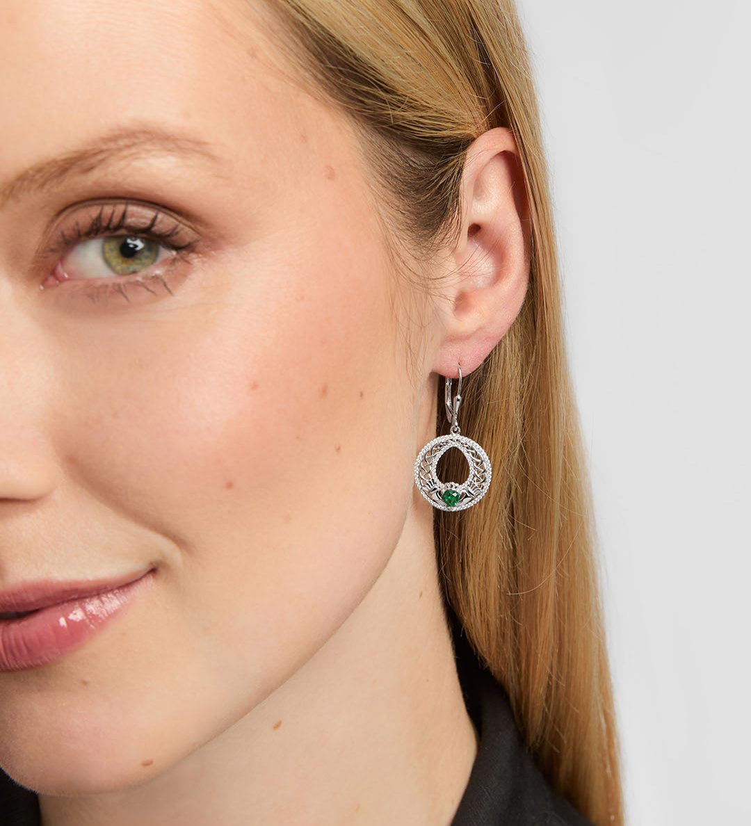 A close-up of a woman's ear and side profile wearing a silver Celtic Claddagh Drop earrings with a circular halo design and a green teardrop-shaped stone at the centre.