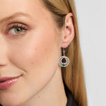 A close-up of a woman's ear and side profile wearing a silver Celtic Claddagh Drop earrings with a circular halo design and a green teardrop-shaped stone at the centre.