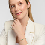 A female model wearing a white blazer and a delicate gold Claddagh bracelet with emerald gemstones, posing against a light grey background.