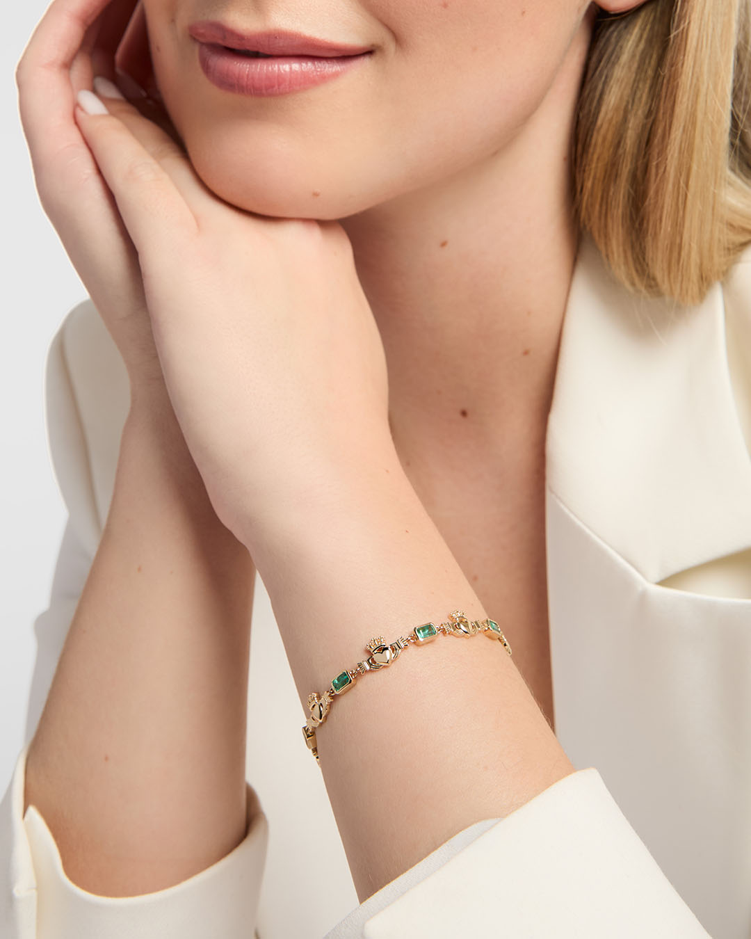 A close-up lifestyle shot of a woman wearing a delicate gold Claddagh bracelet with emerald gemstones on her wrist, paired with a white satin blouse.