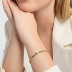 A close-up lifestyle shot of a woman wearing a delicate gold Claddagh bracelet with emerald gemstones on her wrist, paired with a white satin blouse.