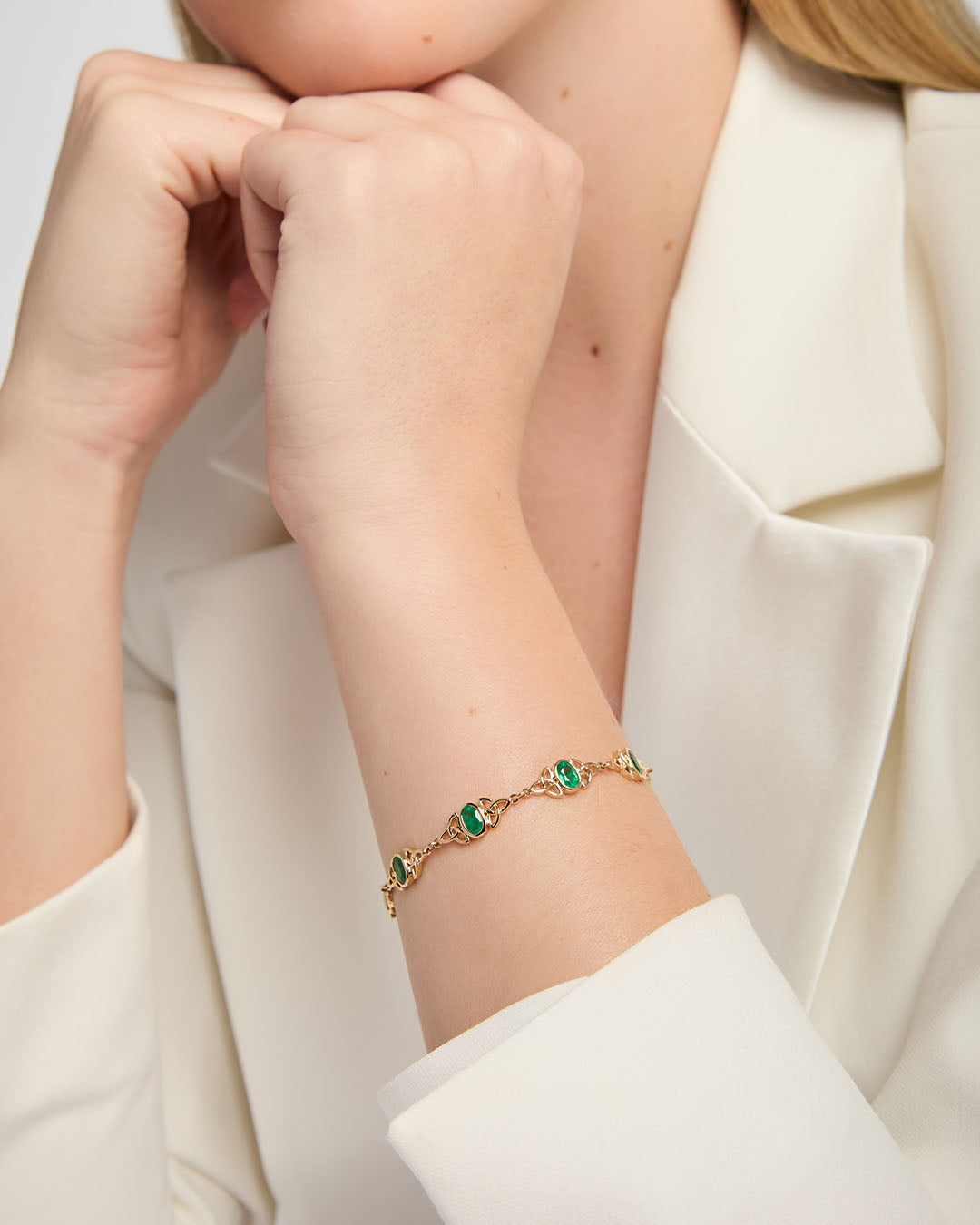 A close-up of a model wearing gold Celtic bracelet with oval real emerald gemstones and decorative knotwork links,