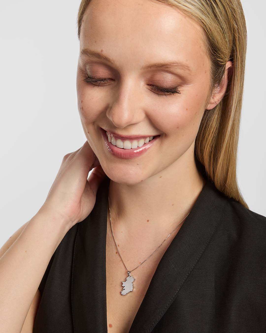 a close-up portrait of a female wearing a dark top and a thin chain necklace. The necklace features a large shiny Map of Ireland pendant