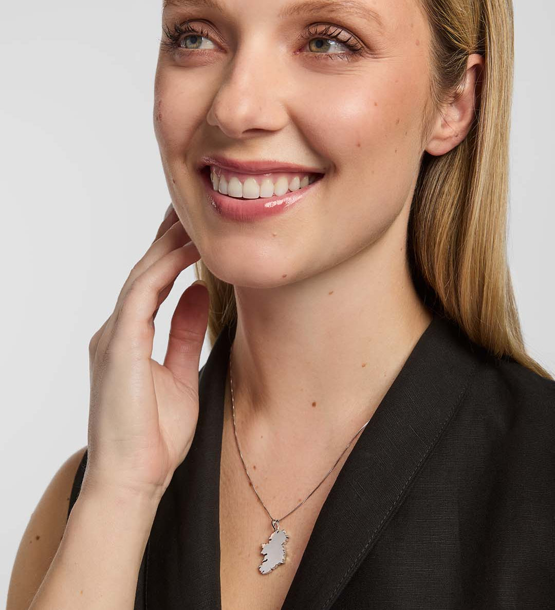 a close-up portrait of a female wearing a dark top and a thin chain necklace. The necklace features a large pendant shaped like the island of Ireland, positioned at the centre of the chest.