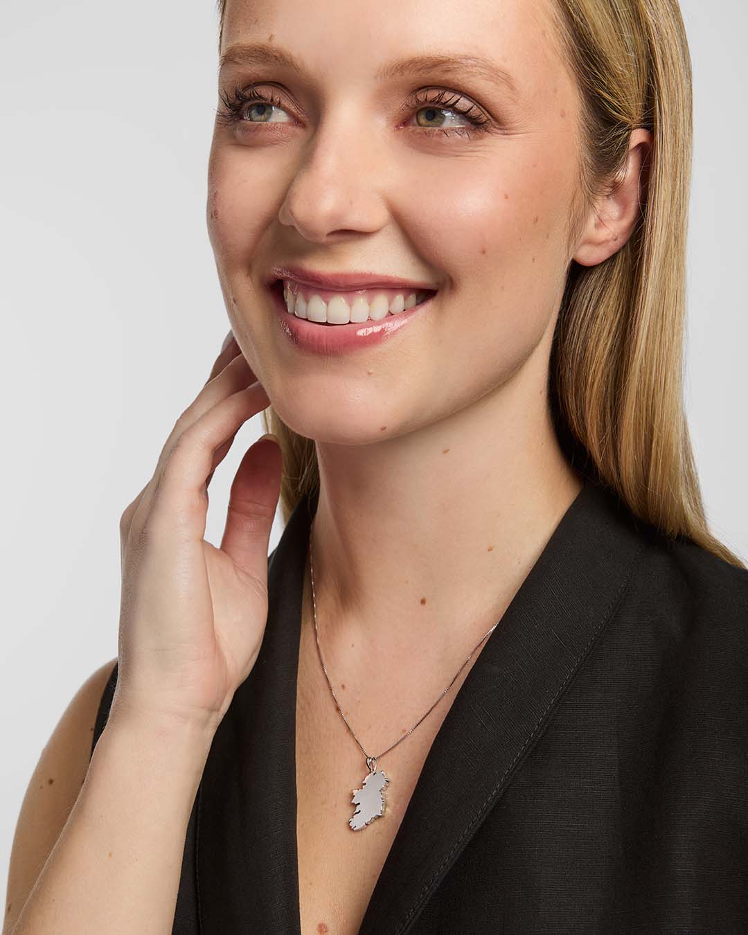 a close-up portrait of a female wearing a dark top and a thin chain necklace. The necklace features a large pendant shaped like the island of Ireland, positioned at the centre of the chest.