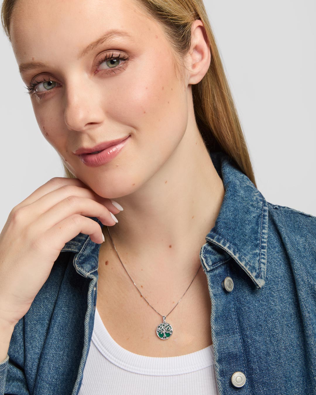 A close-up of a woman wearing a delicate silver Celtic necklace with a circular tree of life pendant featuring green malachite accents, styled with a blue denim shirt over a white top.