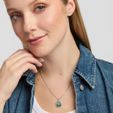A close-up of a woman wearing a delicate silver Celtic necklace with a circular tree of life pendant featuring green malachite accents, styled with a blue denim shirt over a white top.