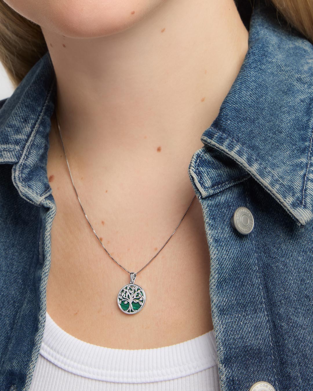 A close-up of a person wearing a silver tree of life pendant necklace with a green malachite background, styled with a blue denim jacket over a white top.