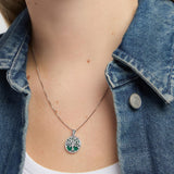 A close-up of a person wearing a silver tree of life pendant necklace with a green malachite background, styled with a blue denim jacket over a white top.