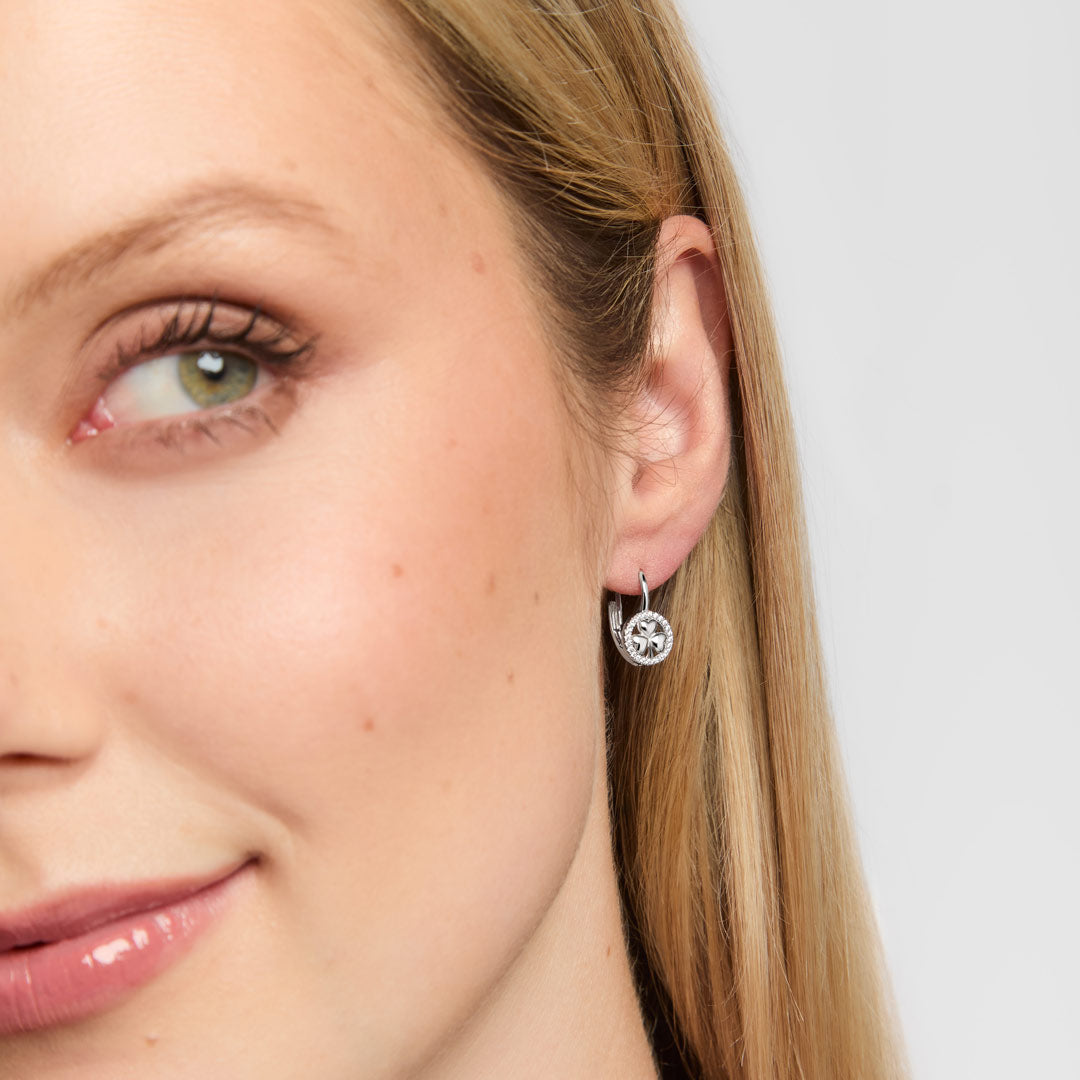 A close-up of a woman' wearing a small silver shamrock hoop earring surrounded by sparkling clear stones, against a light grey background.