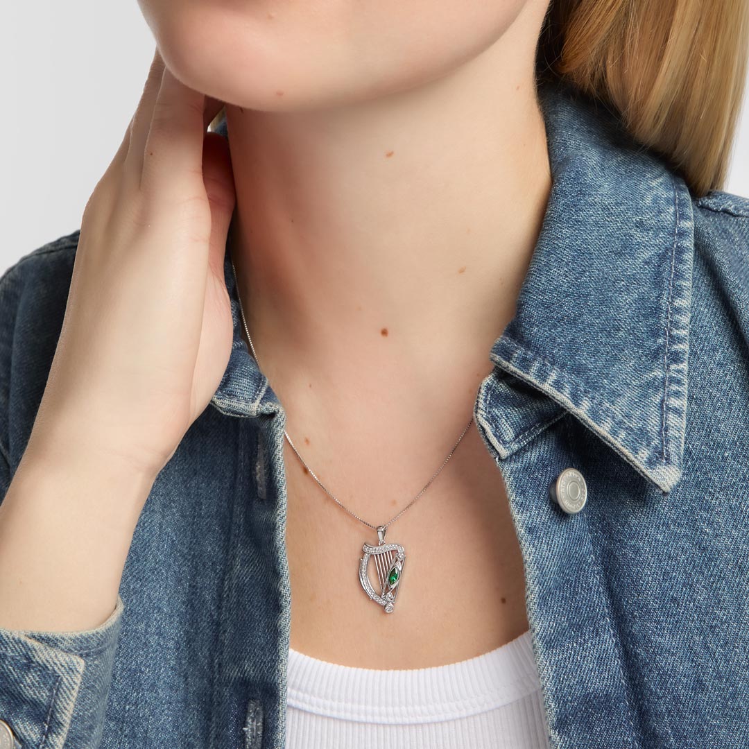 A close-up of a person wearing a silver harp pendant necklace with a small green stone accent, styled with a blue denim shirt over a white top.