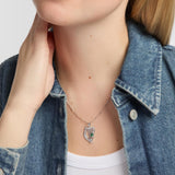 A close-up of a person wearing a silver harp pendant necklace with a small green stone accent, styled with a blue denim shirt over a white top.