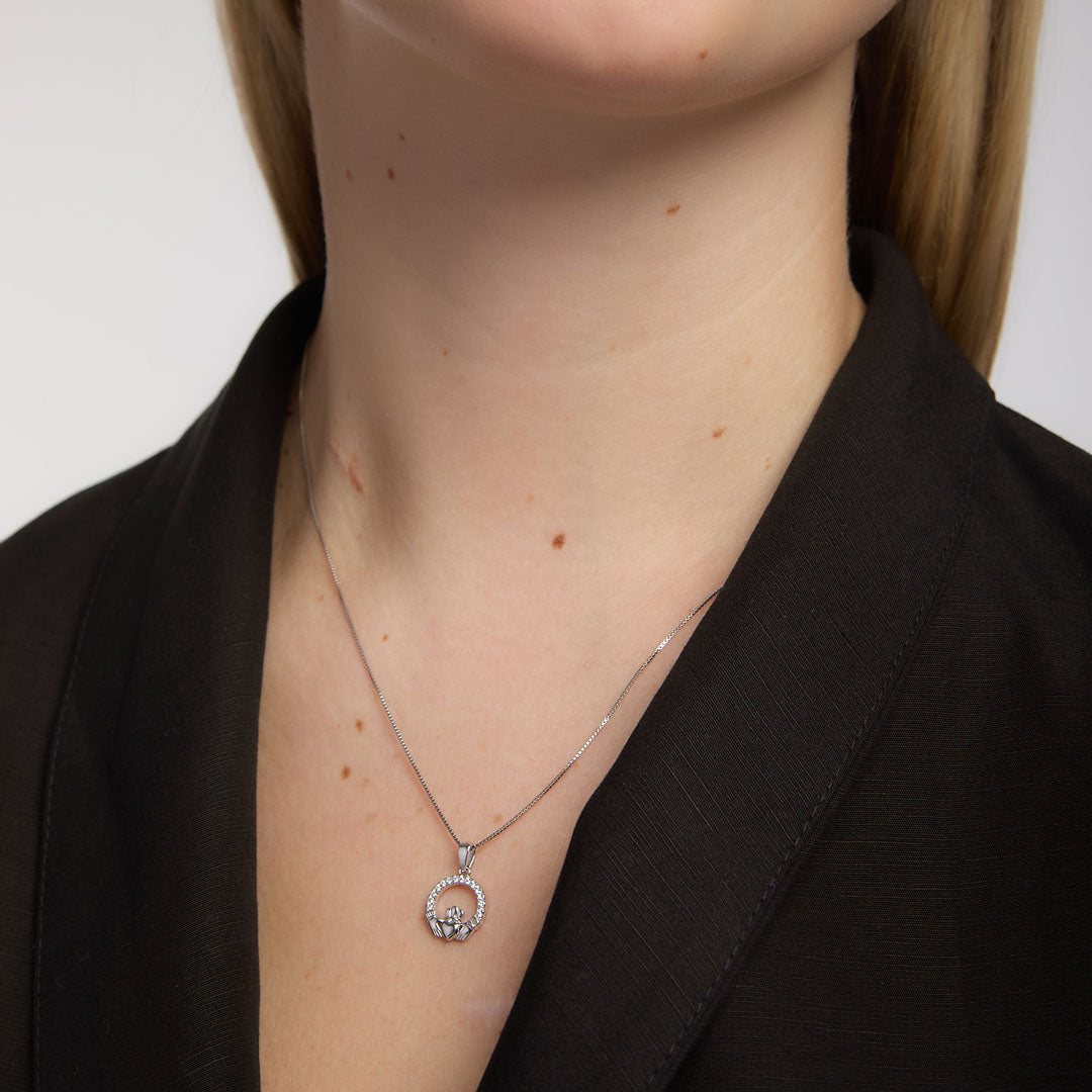 A close-up of a model wearing a delicate silver chain necklace featuring Claddagh design with a circular pendant studded with small clear crystals, styled with a black blazer.