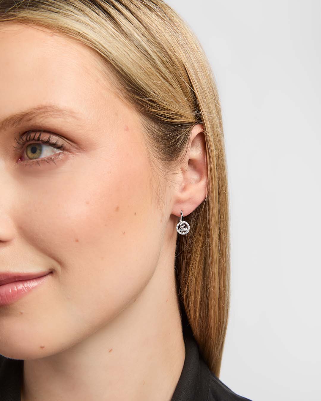 A close-up side profile of a woman wearing a small silver hoop earring with a Trinity Knot centre surrounded by sparkling clear stones.